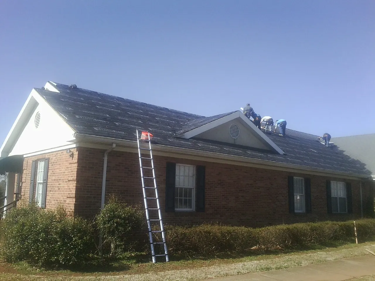 Crew tearing shingles off a brick ranch roof with a ladder set — Emergency Roof Repair work by our crew in Sugar Hill