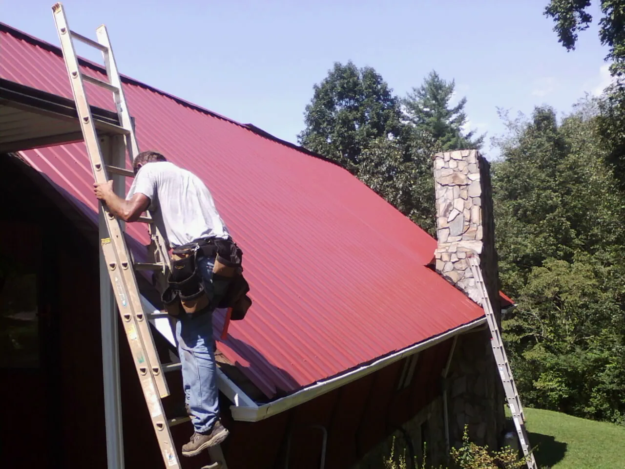 Roofer on a ladder beside a red metal roof and stone chimney — Soffit & Fascia Repair by Roofing Brothers in Sugar Hill