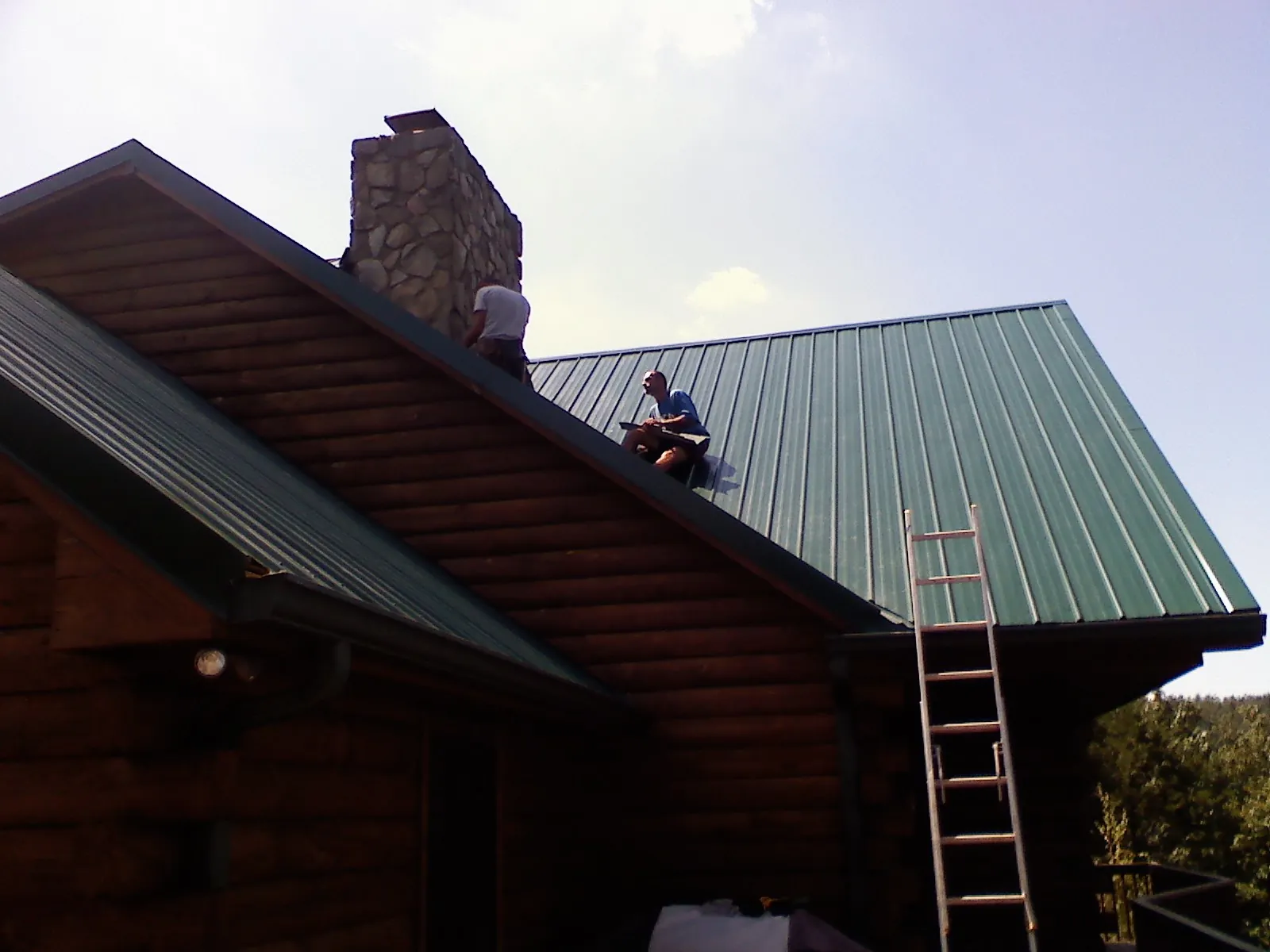Roofers installing green metal panels beside a stone chimney on a log home in Sugar Hill